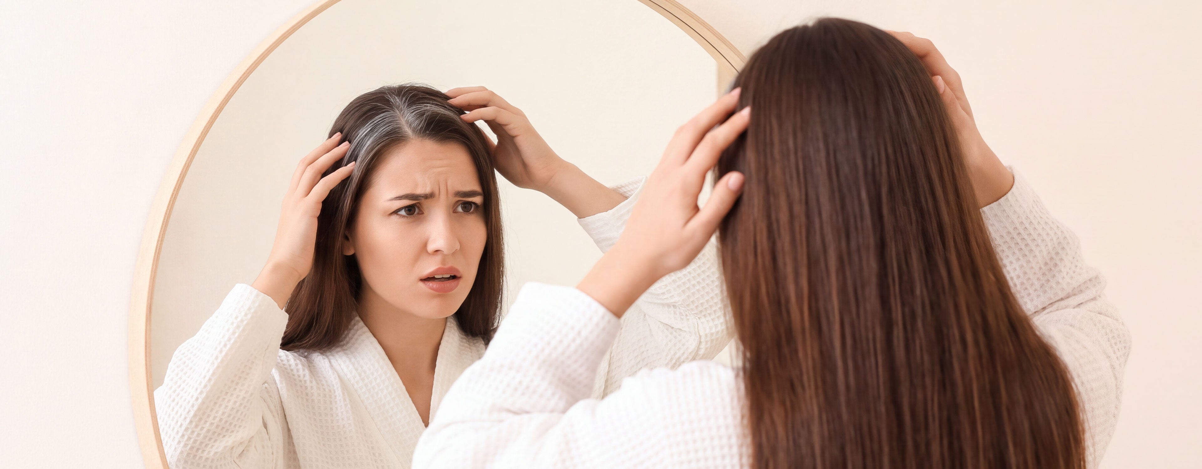 Young woman looks worried as she checks grey roots in the mirror, noticing premature greying in her dark hair